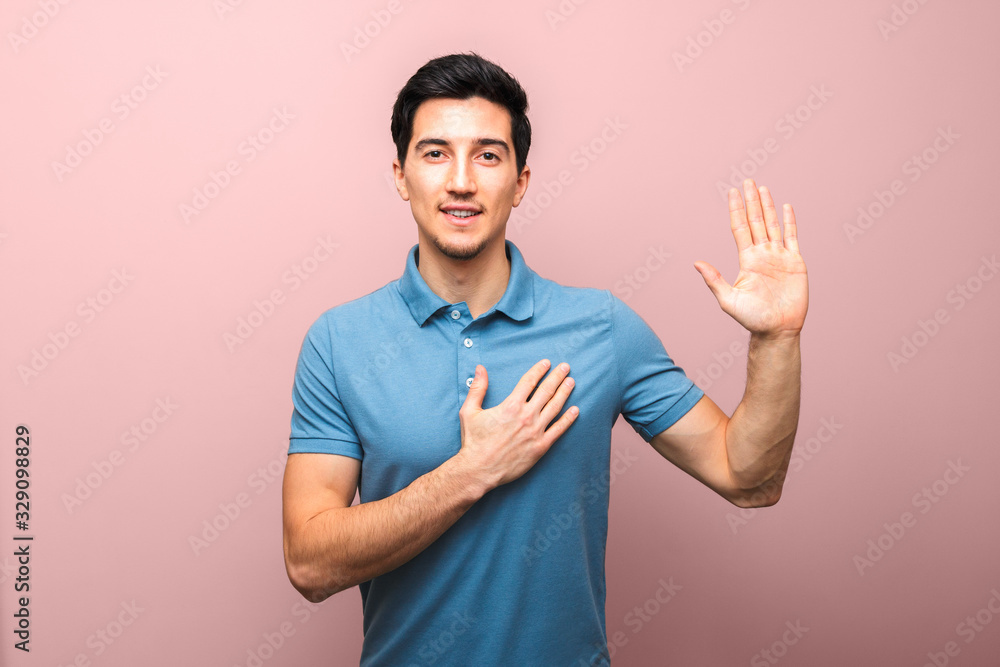 I swear. handsome young man with neutral smile in blue polo shirt with hand on chest giving oath against pink background. studio shoot I swear. handsome young man with neutral smile in blue polo shirt with hand on chest giving oath against pink background. studio shoot