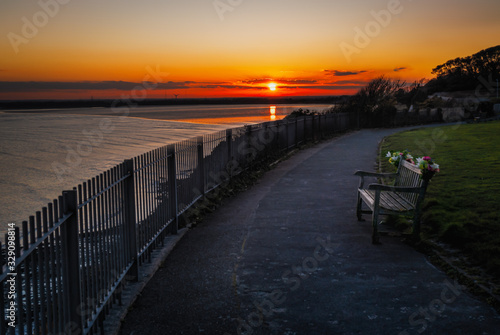 Sunset reflecting in the sea with the cliff top promenade and memorial bench in the foreground in soft focus.