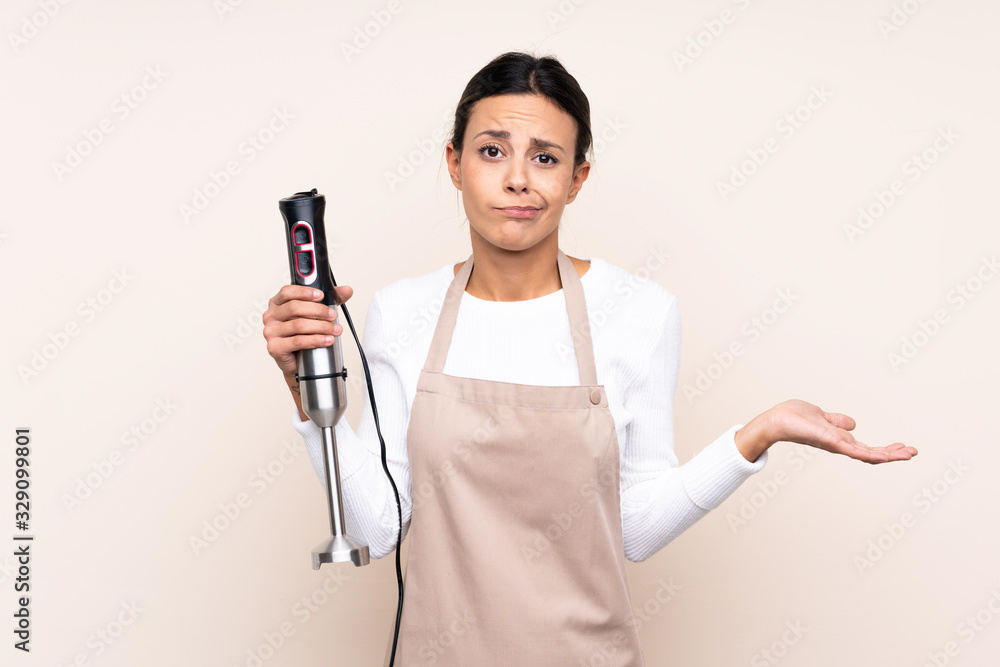 Woman using hand blender over isolated background making doubts gesture while lifting the shoulders