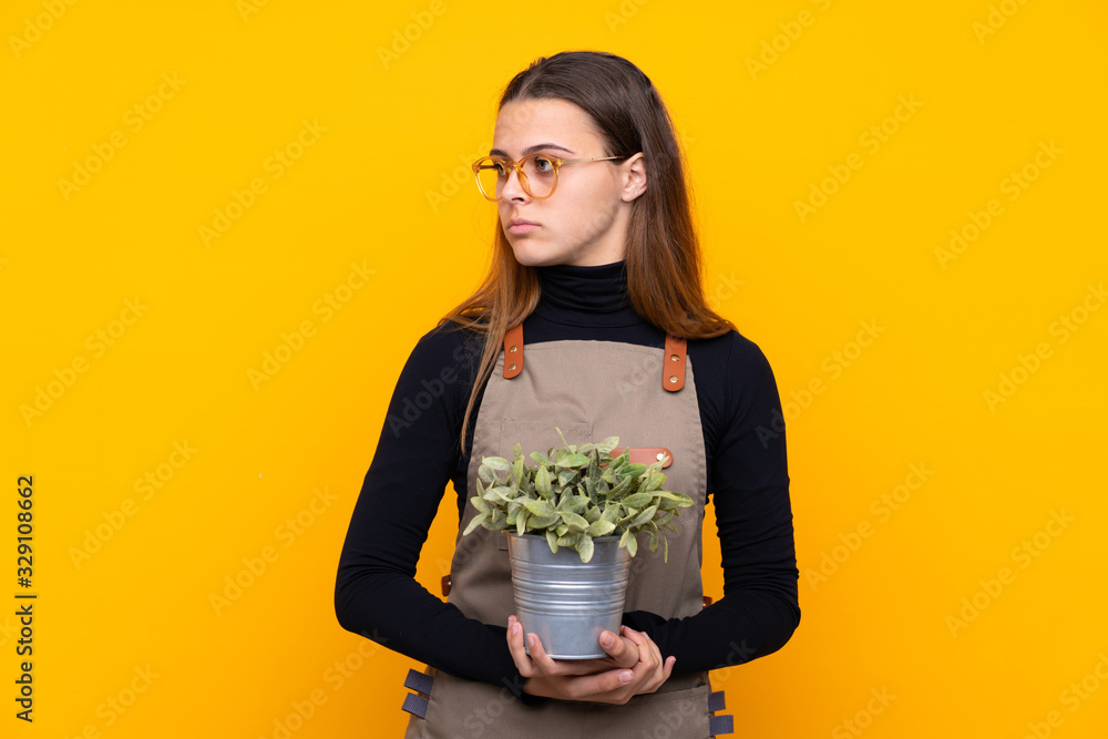 Young gardener girl holding a plant over isolated yellow background looking side Young gardener girl holding a plant over isolated yellow background looking side