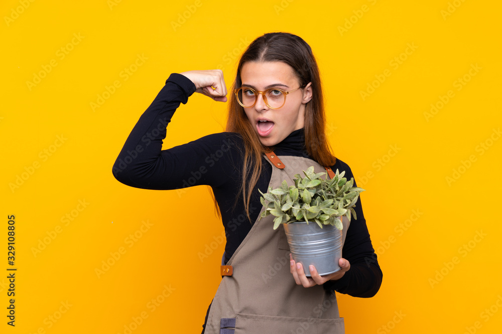 Young gardener girl holding a plant over isolated yellow background making strong gesture Young gardener girl holding a plant over isolated yellow background making strong gesture