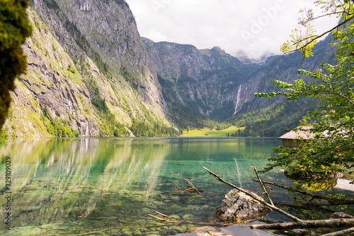 Der Obersee im Berchtesgadener Land ist gerahmt von den Alpen. Ein Wasserfall enspringt im Hintergrund des Bildes. Das Wasser des Bergsees ist klar und die Felswände spiegeln sich im Wasser. 