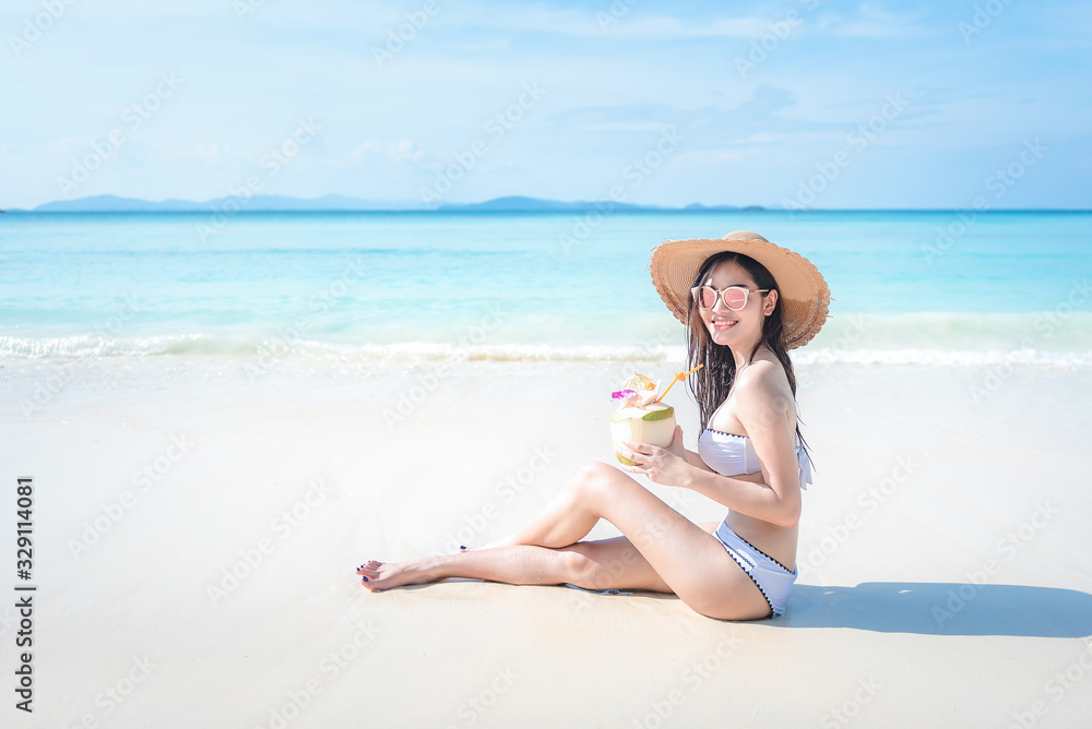 beautiful young tan skin asian woman sitting on the beach wearing bikini ,beach hat, sunglasses smiling with coconut drink. relaxing summer holiday with sunny blue sky, vacation concept