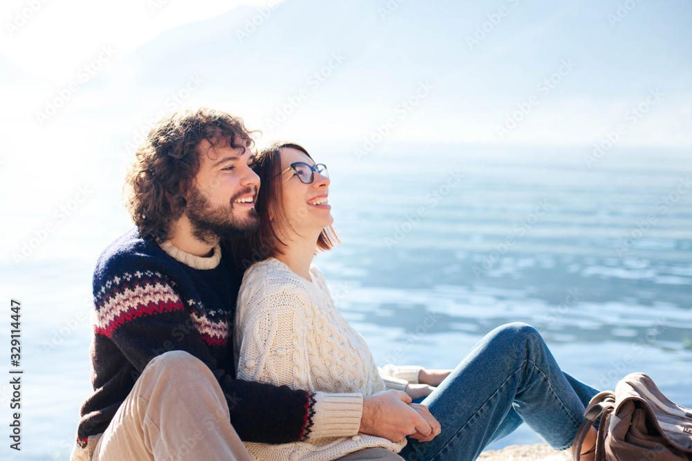 Couple in love hugging and sitting at winter sea beach outdoors. Young travelers smiling and laughing. Happy man and woman have fun. Romantic lifestyle moment.