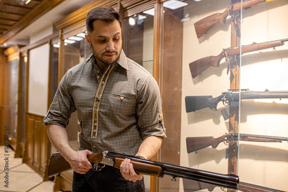 portrait of young caucasian hunter man going to buy new weapon in guns store, he hold rifle in hands, try to use it. indoors, around showcases with rifles portrait of young caucasian hunter man going to buy new weapon in guns store, he hold rifle in hands, try to use it. indoors, around showcases with rifles