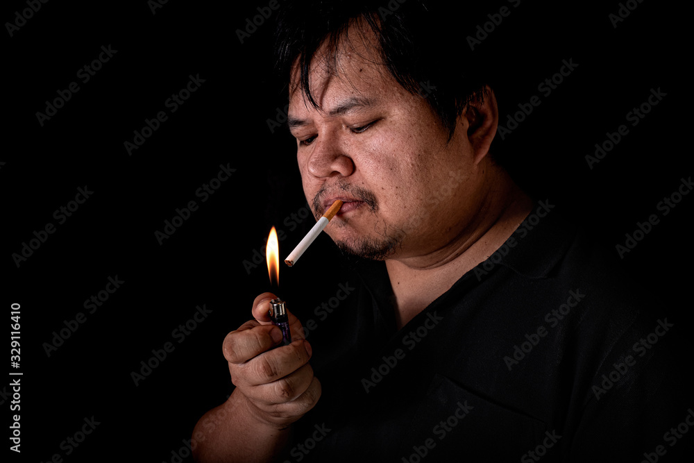 Fototapeta premium The fat man lighting up a joint with lighter smoking at the black background, Image of cigarette smoke spread in hand