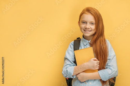 portrait of adorable smiling caucasian school girl with bag and book isolated over yellow background. 6 years old girl dream to go at school