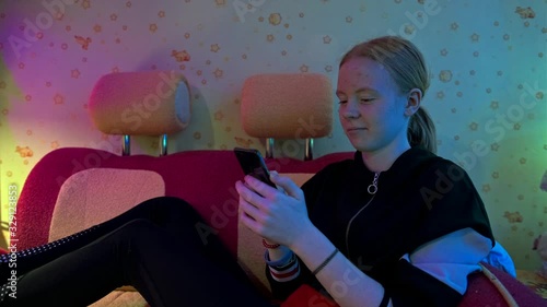 cheerful schoolgirl with snow-white skin plays on digital device and smiles sitting on red couch in dim room close view photo