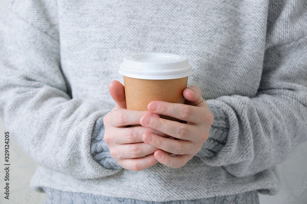 A paper brown cup of coffee in the hand of a caucasian girl in a gray sweater. A paper brown cup of coffee in the hand of a caucasian girl in a gray sweater.