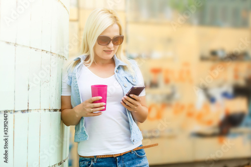Cheerful young woman using her phone in the sunny city street and drinking take away coffee in pink cup.
