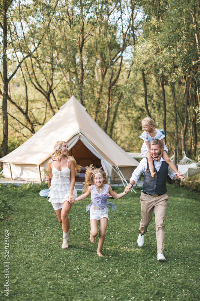 Fototapeta premium Happy Caucasian family, father mother and two little daughters, having fun outdoors in park or camping, enjoying time and laughing together, holding hands and running. Love and parenthood concept