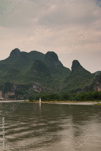 Guilin, China - May 10, 2010: Along Li River. Long shot of white and red navigation beacon standing at edge of water with forest covered karst mountains in back under rain cloudscape. © Klodien