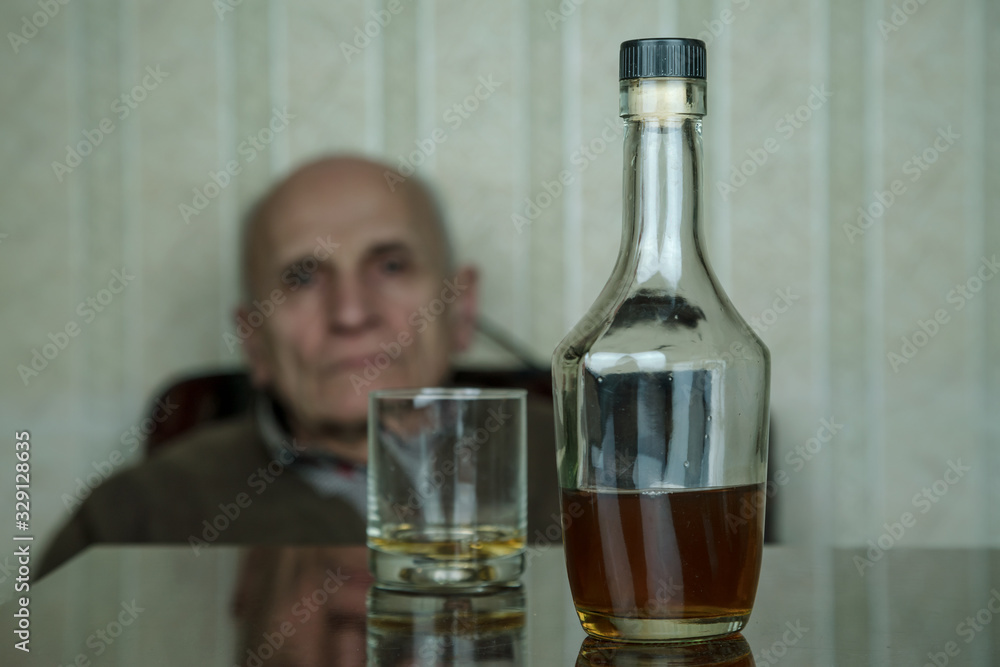 blurry old man looks at bottle and glass with cognac on brown wooden table at home blurry old man looks at bottle and glass with cognac on brown wooden table at home