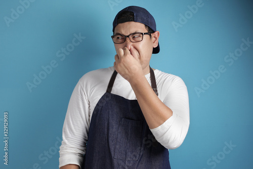 Portrait of funny young male Asian chef or waiter closing his nose, smells something bad