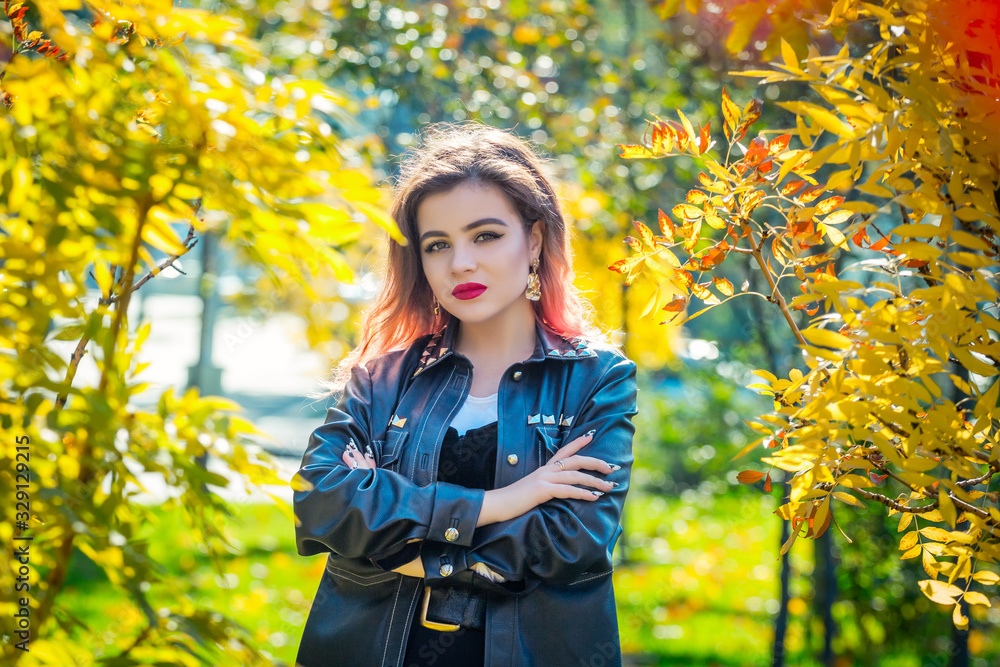 Portrait of beautiful stylish young woman with bright make-up, wearing black leather jacket, standing in autumn park near the tree with bright yellow foliage. Portrait of beautiful stylish young woman with bright make-up, wearing black leather jacket, standing in autumn park near the tree with bright yellow foliage.