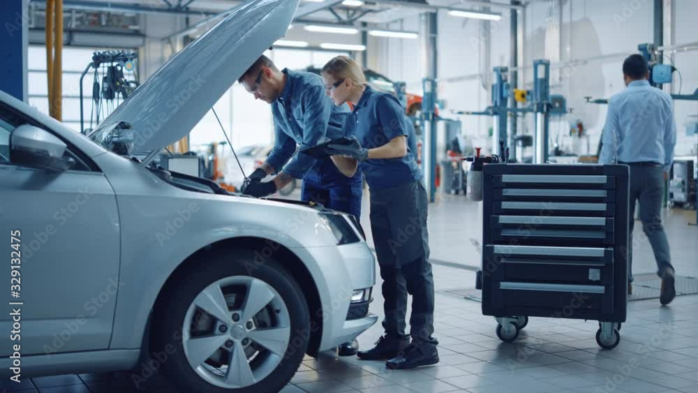 Two Mechanics in a Service are Inspecting a Car After They Got the Diagnostics Results. Female Specialist is Comparing the Data on a Tablet Computer. Repairman is Using a Ratchet to Repair the Faults. Two Mechanics in a Service are Inspecting a Car After They Got the Diagnostics Results. Female Specialist is Comparing the Data on a Tablet Computer. Repairman is Using a Ratchet to Repair the Faults.