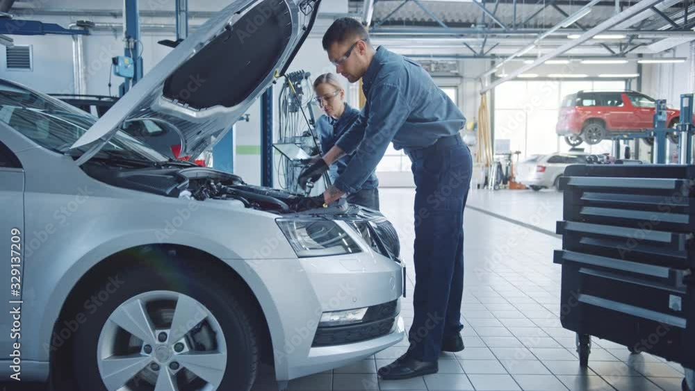 Two Mechanics in a Service are Inspecting a Car After They Got the Diagnostics Results. Female Specialist is Comparing the Data on a Tablet Computer. Repairman is Using a Ratchet to Repair the Faults.