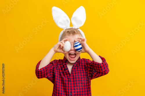 Excited boy in rabbit bunny ears on head on yellow studio background. Cheerful crazy smiling happy child. Easter blue color of the year painting eggs on eyes. Easter and holidays.
