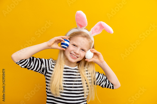 Girl in rabbit bunny ears on head on yellow studio background. Cheerful crazy smiling happy child. Easter blue color of the year painting eggs on eyes. Easter and holidays.