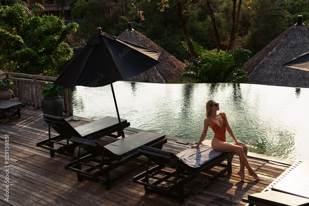 Woman in swimsuit and sunglasses relaxing at sunbed near luxury infinity pool with jungle view