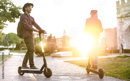 Modern couple using electric scooter in city park - Milenial students riding new ecological mean of transport - Green eco energy concept with zero emission - Bright warm filter with sunshine halo photo