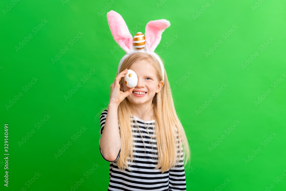 Girl in rabbit bunny ears on head on green studio background. Cheerful crazy smiling happy child. Easter painting eggs on eyes. Easter and holidays