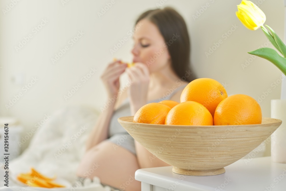 Bowl with oranges in focus, pregnant young woman eating slices of oranges sitting in bed Bowl with oranges in focus, pregnant young woman eating slices of oranges sitting in bed