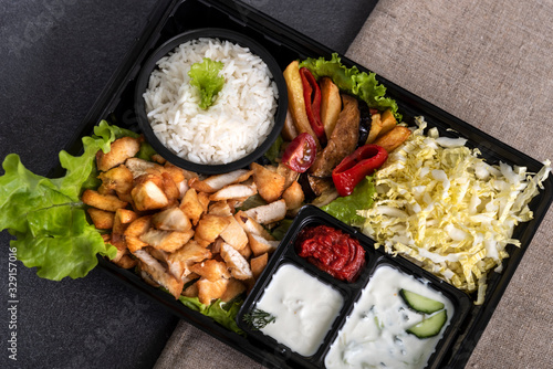 Traditional doner kebab in a black plastic container, sour cream, white boiled rice, roast meat, chiken, sauces, salad, cabbage and roasted vegetables on grey background