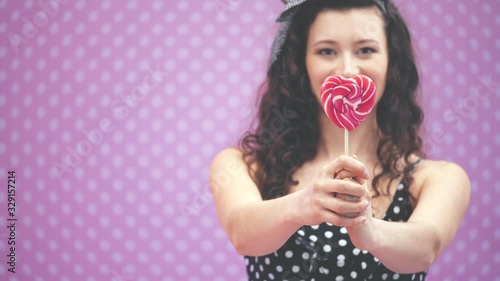 Lovely young curly girl standing, extending red lollipop on the stick to the camera. Blurred background. Focus on the candy on the forefront. photo