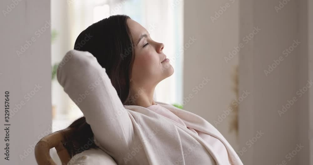 Relaxed serene millennial woman breathing fresh air with eyes closed resting on comfortable chair at home. Calm lady enjoys lounge and nap holds hands behind head. No stress concept. Closeup side view Relaxed serene millennial woman breathing fresh air with eyes closed resting on comfortable chair at home. Calm lady enjoys lounge and nap holds hands behind head. No stress concept. Closeup side view