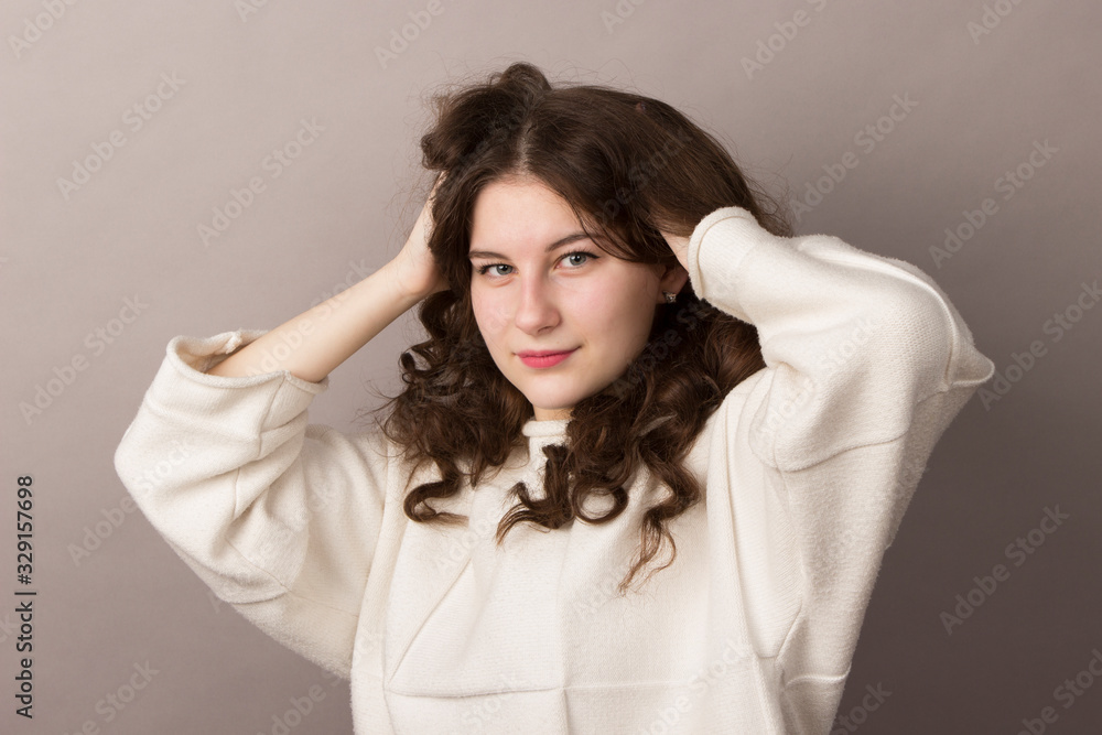Naklejka premium Beautiful young brunette teenager girl with wavy long curly thick hair in a white sweater on a gray background. Waist photo