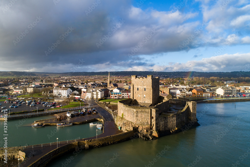 Medieval Norman Castle, harbor with boat ramp and wave breaker in Carrickfergus near Belfast, Northern Ireland, UK. Aerial view in sunset light in winter. Town and stormy clouds in the background
