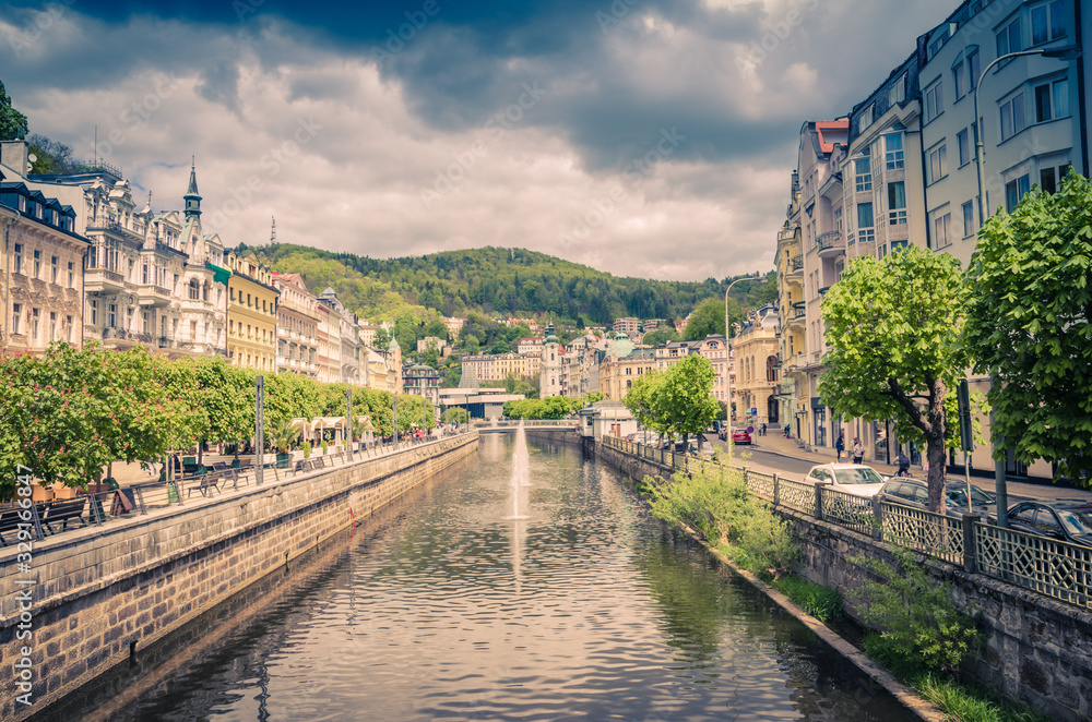 Karlovy Vary (Carlsbad) historical city centre with Tepla river central embankment, colorful beautiful buildings, Slavkov Forest hills with green trees background, West Bohemia, Czech Republic
