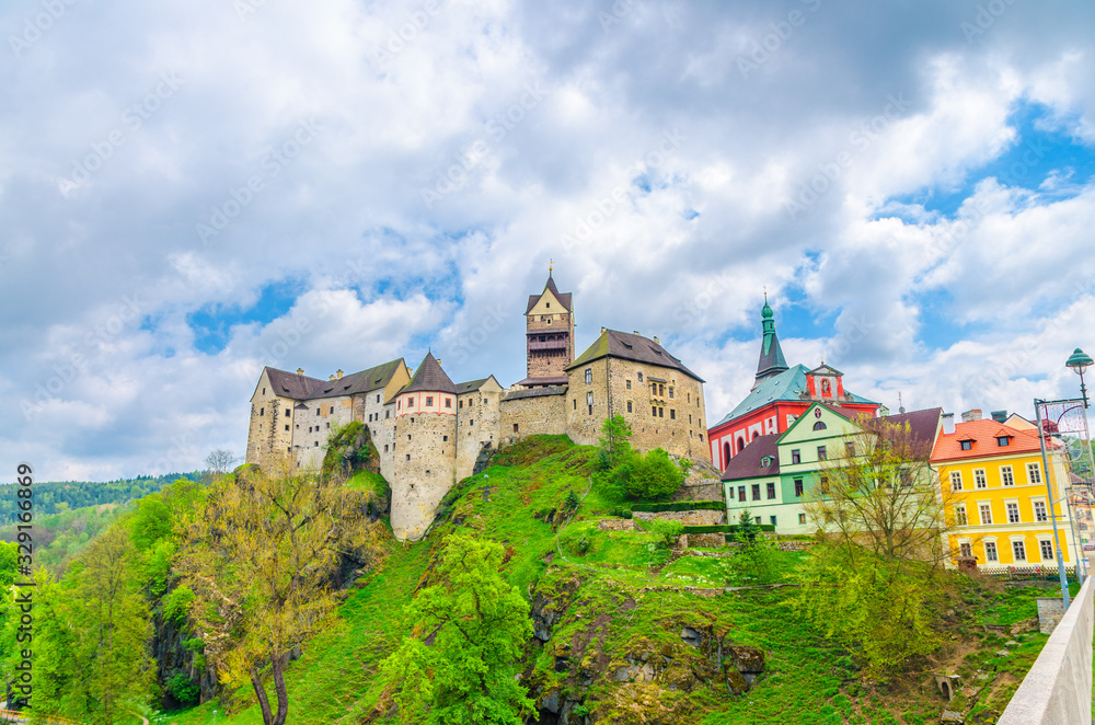 Loket Castle Hrad Loket gothic style building on massive rock and colorful buildings in Loket town, green trees and hills, blue cloudy sky background, Karlovy Vary Region, West Bohemia, Czech Republic
