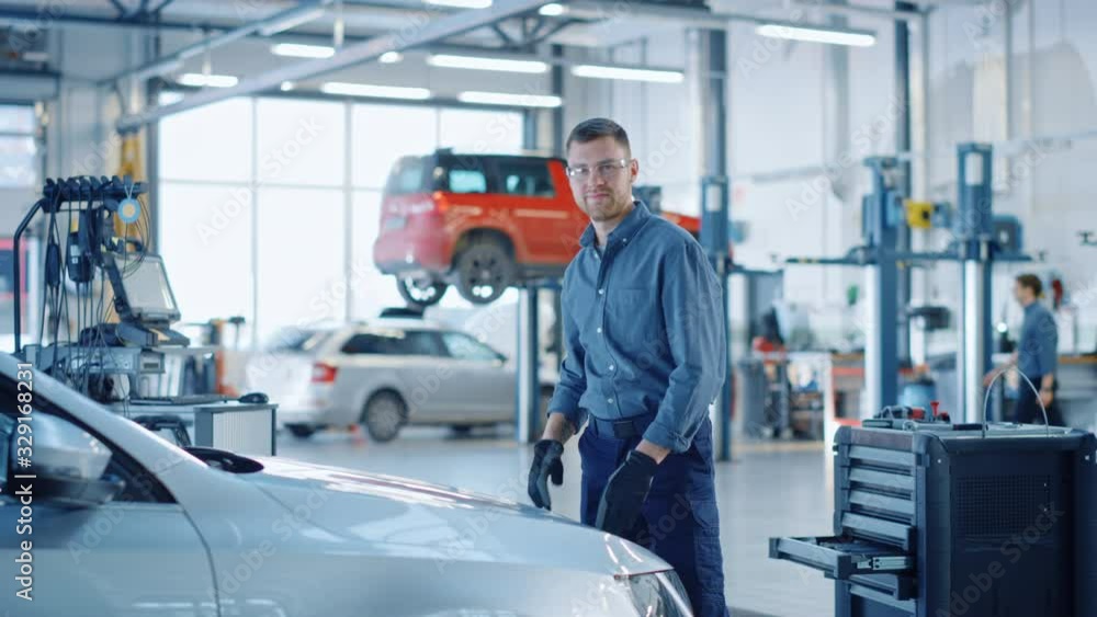 Handsome Professional Car Mechanic is Working on a Vehicle in a Service. Happy Repairman is Closing the Hood. Specialist is Wearing Safety Glasses. He Looks at a Camera and Smiles. Handsome Professional Car Mechanic is Working on a Vehicle in a Service. Happy Repairman is Closing the Hood. Specialist is Wearing Safety Glasses. He Looks at a Camera and Smiles.