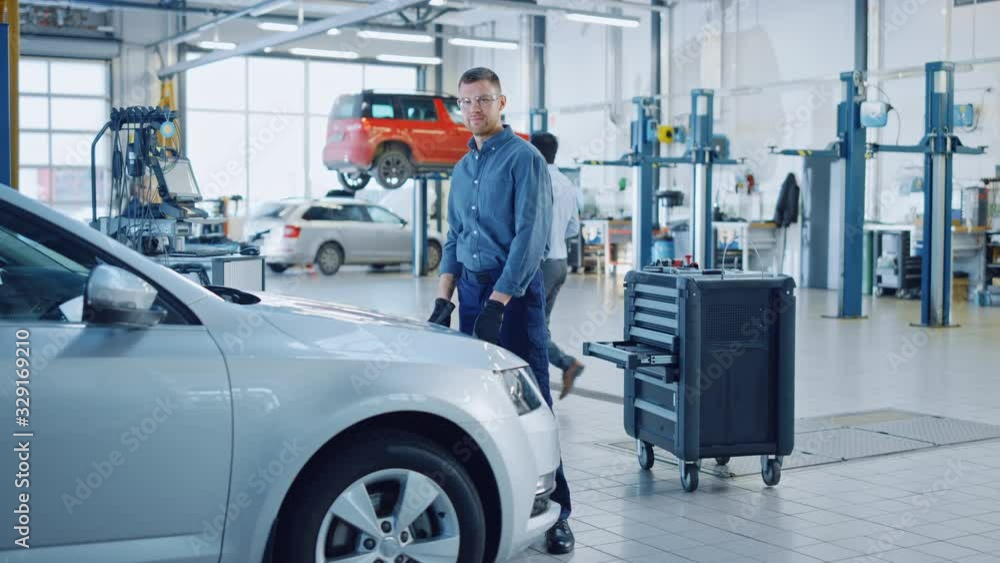 Handsome Professional Car Mechanic is Working on a Vehicle in a Service. Happy Repairman is Closing the Hood. Specialist is Wearing Safety Glasses. He Looks at a Camera and Smiles. Handsome Professional Car Mechanic is Working on a Vehicle in a Service. Happy Repairman is Closing the Hood. Specialist is Wearing Safety Glasses. He Looks at a Camera and Smiles.
