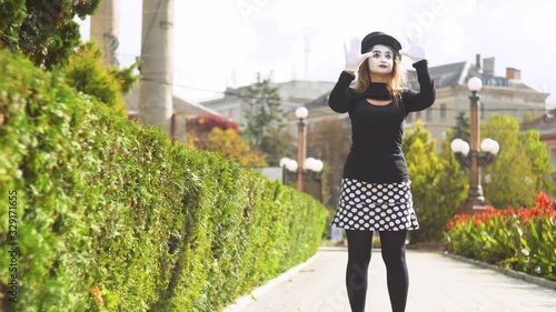 Young funny female mime behind an invisible glass box or wall. Mime on the park shows pantomime