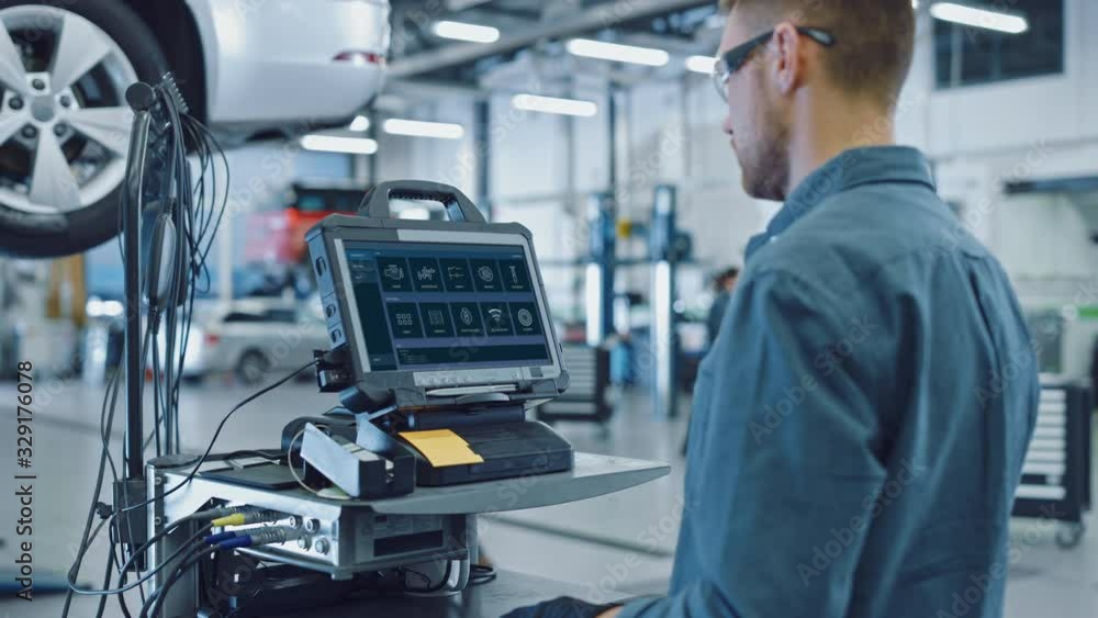 Car Service Manager or Mechanic is Running an Interactive Diagnostics Software on an Advanced Computer. Specialist Inspecting the Vehicle in Order to Find Broken Components and Errors in Data Logs. Car Service Manager or Mechanic is Running an Interactive Diagnostics Software on an Advanced Computer. Specialist Inspecting the Vehicle in Order to Find Broken Components and Errors in Data Logs.