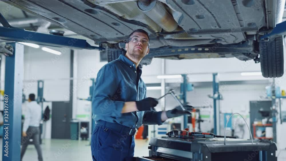 Handsome Professional Car Mechanic is Working Under a Vehicle on a Lift in Service. Repairman is Using a LED lamp and Takes a Ratchet. Specialist is Wearing Safety Glasses. Modern Workshop. Handsome Professional Car Mechanic is Working Under a Vehicle on a Lift in Service. Repairman is Using a LED lamp and Takes a Ratchet. Specialist is Wearing Safety Glasses. Modern Workshop.