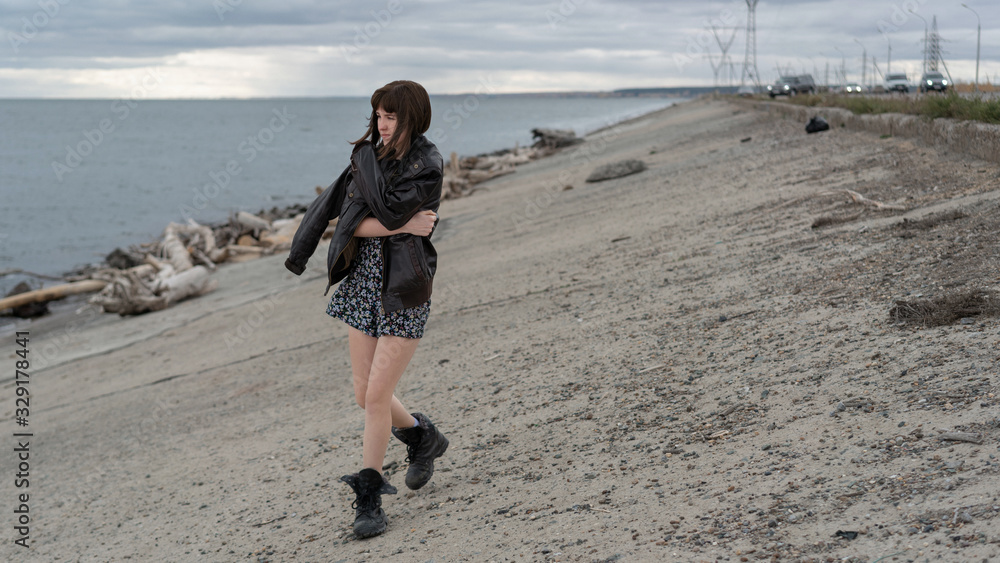 A young girl in a dress, boots and leather jacket walks along the water.