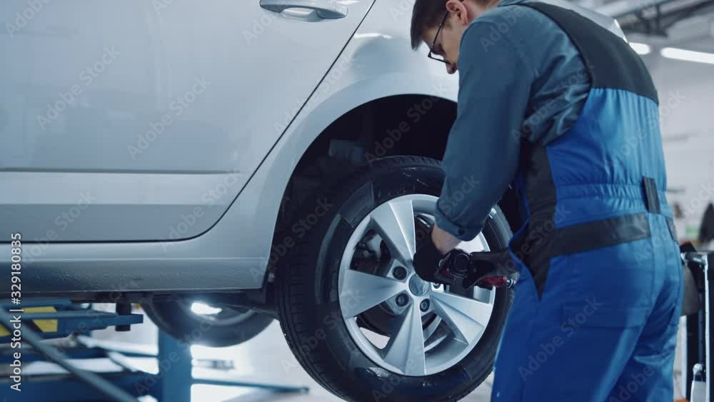 Mechanic in Blue Overalls is Unscrewing Lug Nuts with a Pneumatic Impact Wrench. Repairman Works in a Modern Clean Car Service. Specialists Removes the Wheel in Order to Fix a Component on a Vehicle. Mechanic in Blue Overalls is Unscrewing Lug Nuts with a Pneumatic Impact Wrench. Repairman Works in a Modern Clean Car Service. Specialists Removes the Wheel in Order to Fix a Component on a Vehicle.