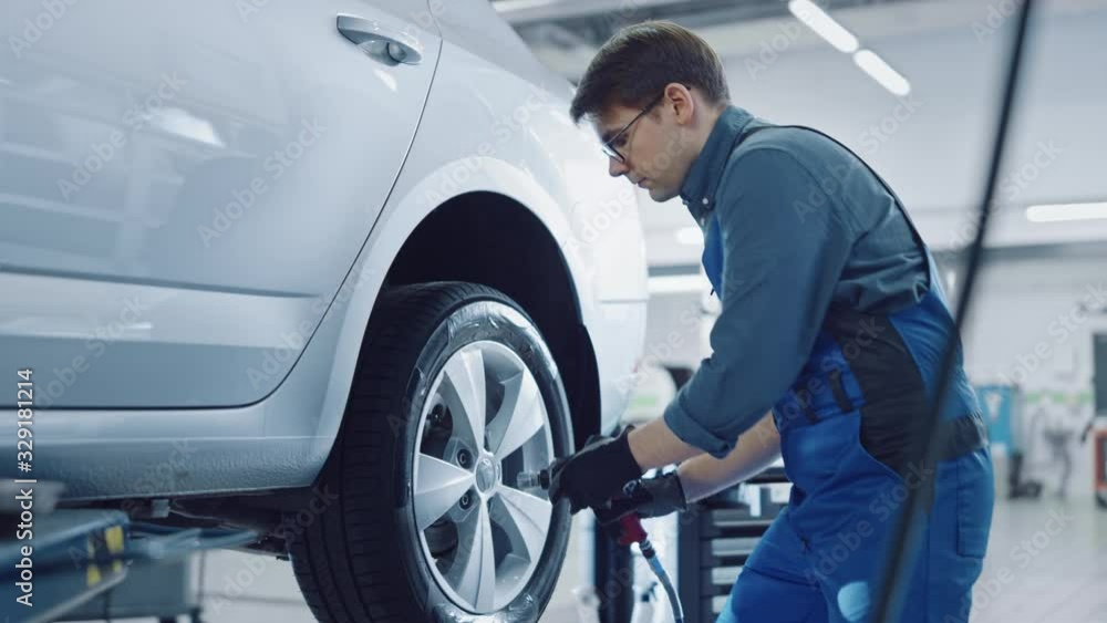 Mechanic in Blue Overalls is Unscrewing Lug Nuts with a Pneumatic Impact Wrench. Repairman Works in a Modern Clean Car Service. Specialists Removes the Wheel in Order to Fix a Component on a Vehicle. Mechanic in Blue Overalls is Unscrewing Lug Nuts with a Pneumatic Impact Wrench. Repairman Works in a Modern Clean Car Service. Specialists Removes the Wheel in Order to Fix a Component on a Vehicle.