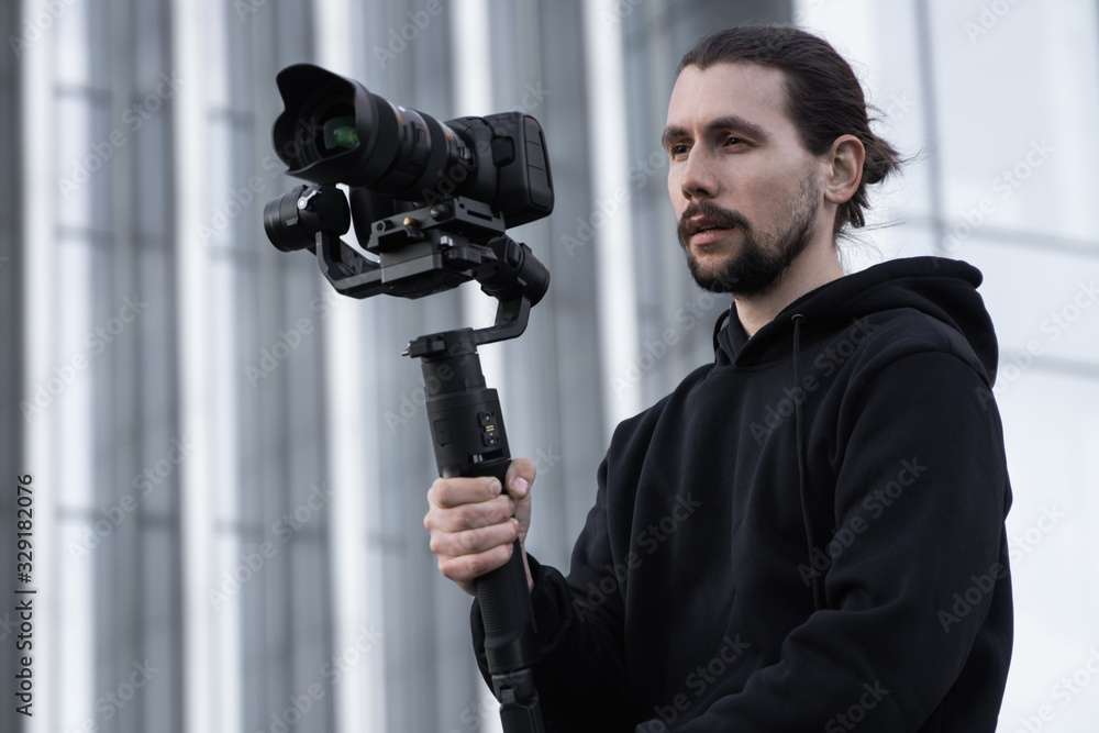 Young Professional videographer holding professional camera on 3-axis gimbal stabilizer. Pro equipment helps to make high quality video without shaking. Cameraman wearing white hoodie making a videos. Young Professional videographer holding professional camera on 3-axis gimbal stabilizer. Pro equipment helps to make high quality video without shaking. Cameraman wearing white hoodie making a videos.