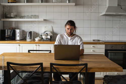 Freelancer working from home on the kitchen and using laptop. Bearded man working with a laptop and reading good news. Handsome successful self entrepreneur sitting and working at his modern home. © Volodymyr