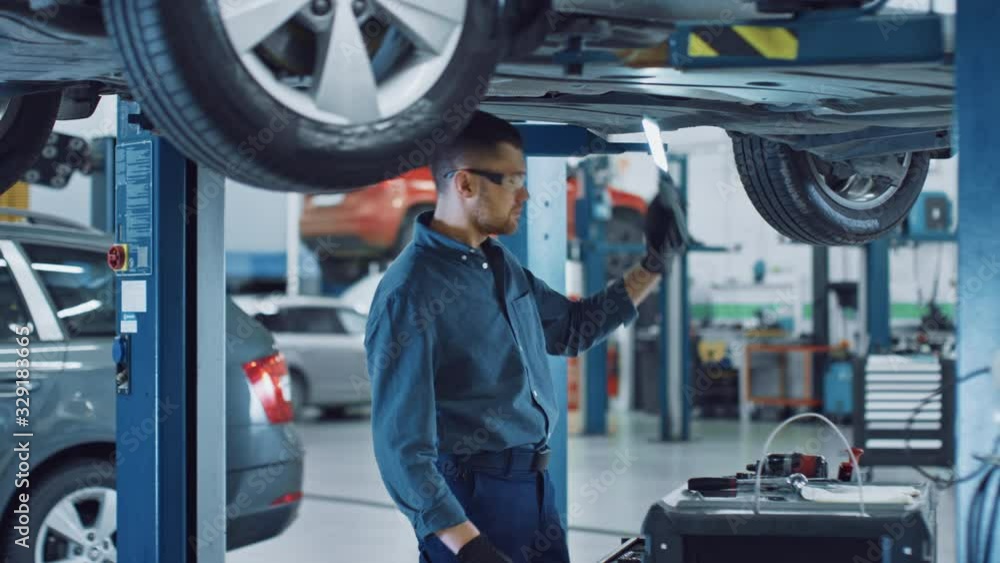 Handsome Professional Car Mechanic is Working Under a Vehicle on a Lift in Service. Repairman is Using a LED lamp and Takes a Ratchet. Specialist is Wearing Safety Glasses. Modern Workshop.