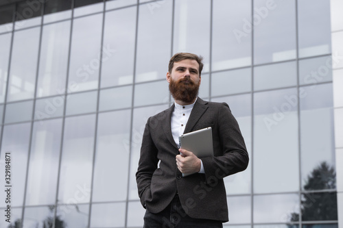 Modern Young bearded Business man standing with a digital tablet. Young hipster businessman holding tablet in hands outdoor. Working online with a tablet while standing outside on an office building.