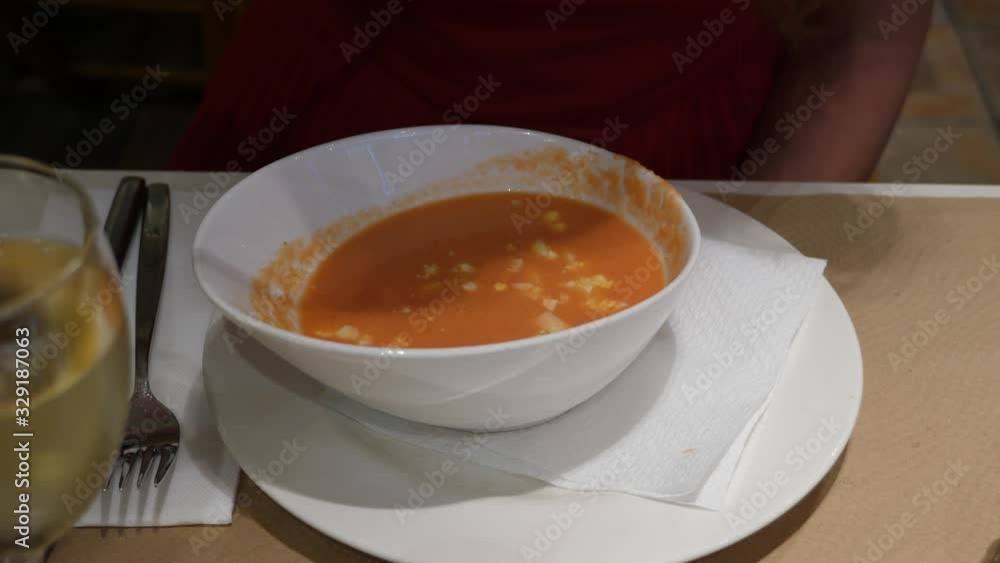 Bowl with traditional Spanish Gazpacho, popular cold soup, woman eat lunch at restaurant. Close up shot of dish standing on white plate on table, glass of white wine seen near Bowl with traditional Spanish Gazpacho, popular cold soup, woman eat lunch at restaurant. Close up shot of dish standing on white plate on table, glass of white wine seen near
