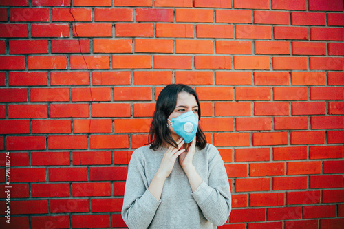 brunette woman stands in a medical mask on her face, which protects against coronovirus and other viruses and diseases against a red brick wall 1