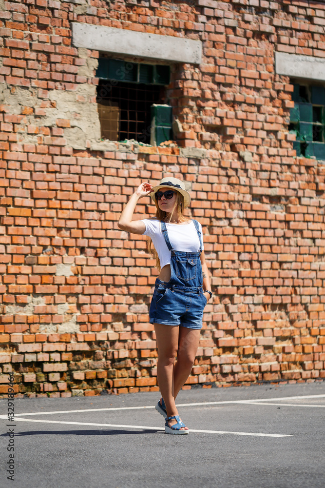 Girl in denim overalls, a white T-shirt and a light hat on a background of a brick building under the bright sun