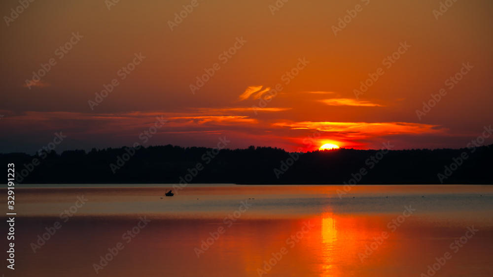 warm summer red-orange sunset over the lake and sun disc behind the forest